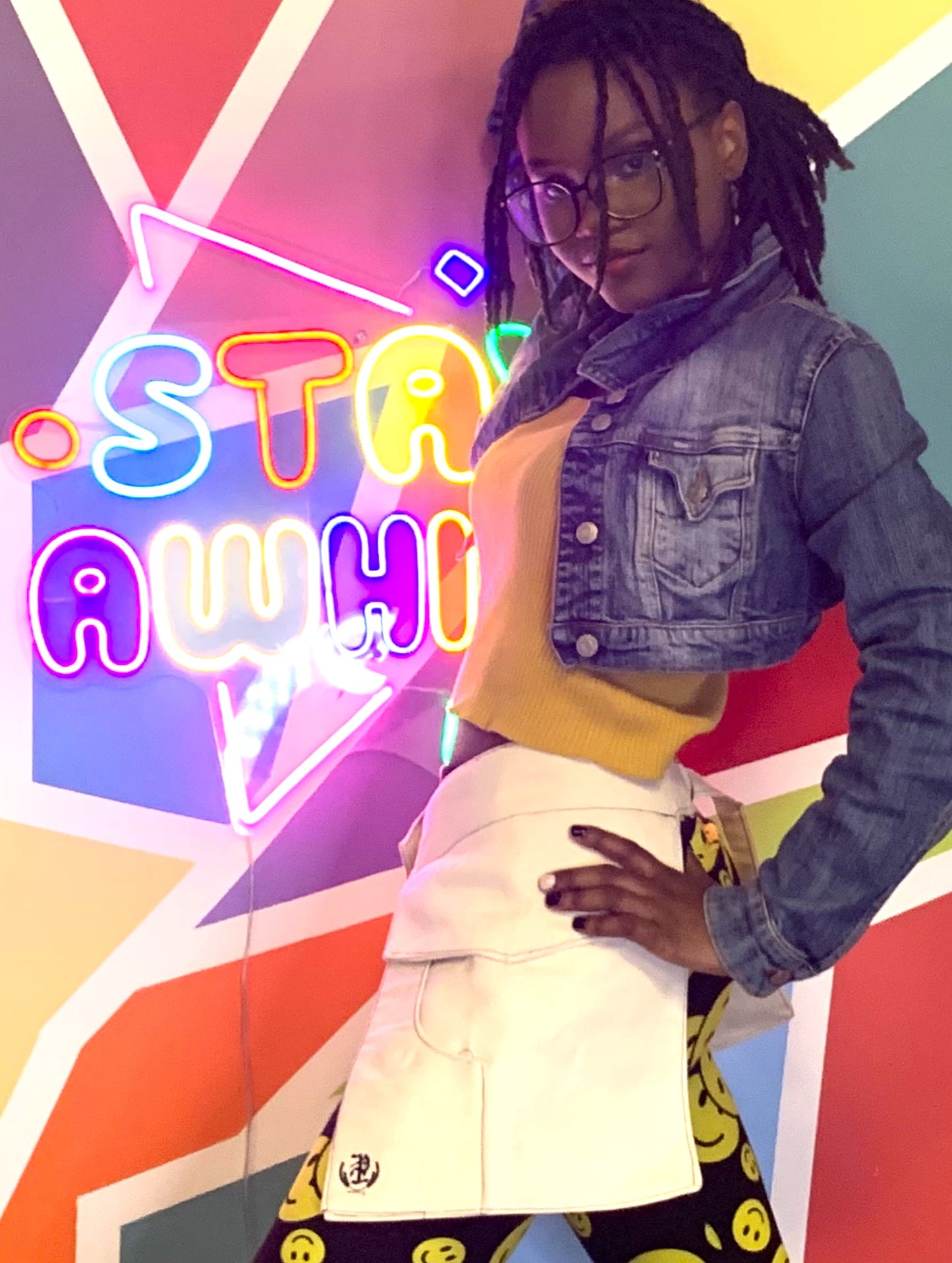 A young woman wearing a pair of off-white pockets with her hands on her hips, standing in front of a neon sign saying "stay awhile" amid a colorful background